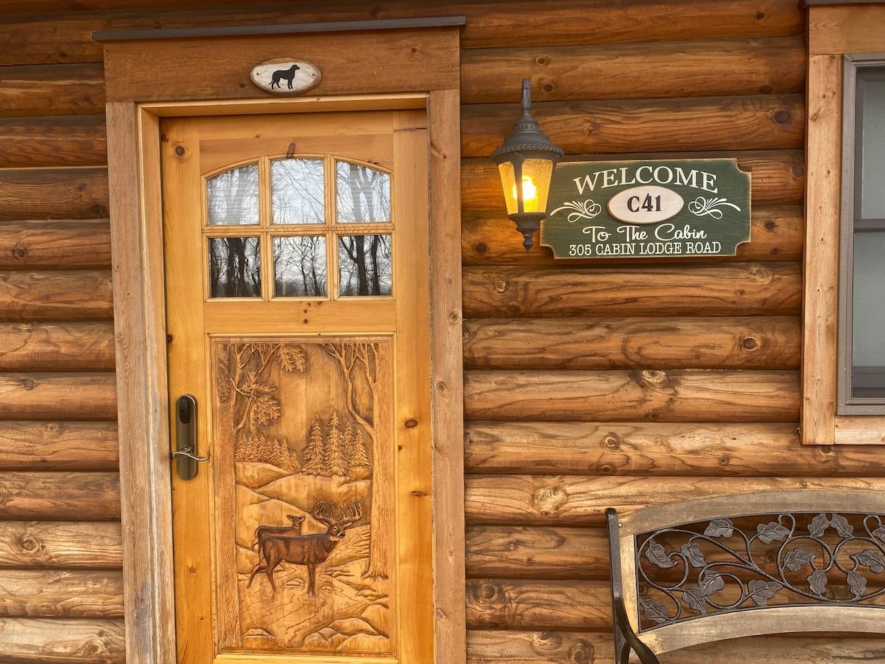 A wooden cabin entrance with a carved door depicting a forest scene, a welcome sign, and a lantern.