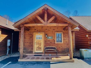 A log cabin with a pitched roof, a wooden front door, a bench, and a welcome sign.