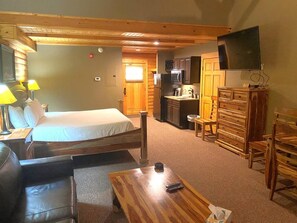 A room with a bed, a wooden table, a television, a kitchen area, and a bathroom.