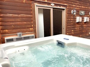 A hot tub in front of a wooden cabin with a sliding door, a welcome sign, and two wall-mounted heaters.