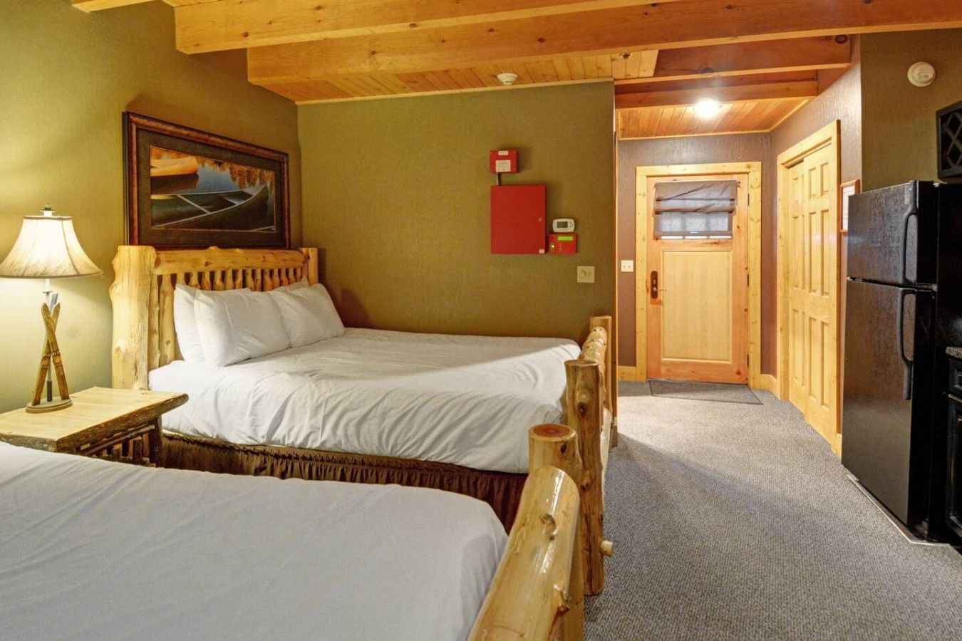 Una habitación con dos camas, cabecera de madera, mesita de noche con lámpara, un cuadro en la pared, detector de incendios rojo y un refrigerador.