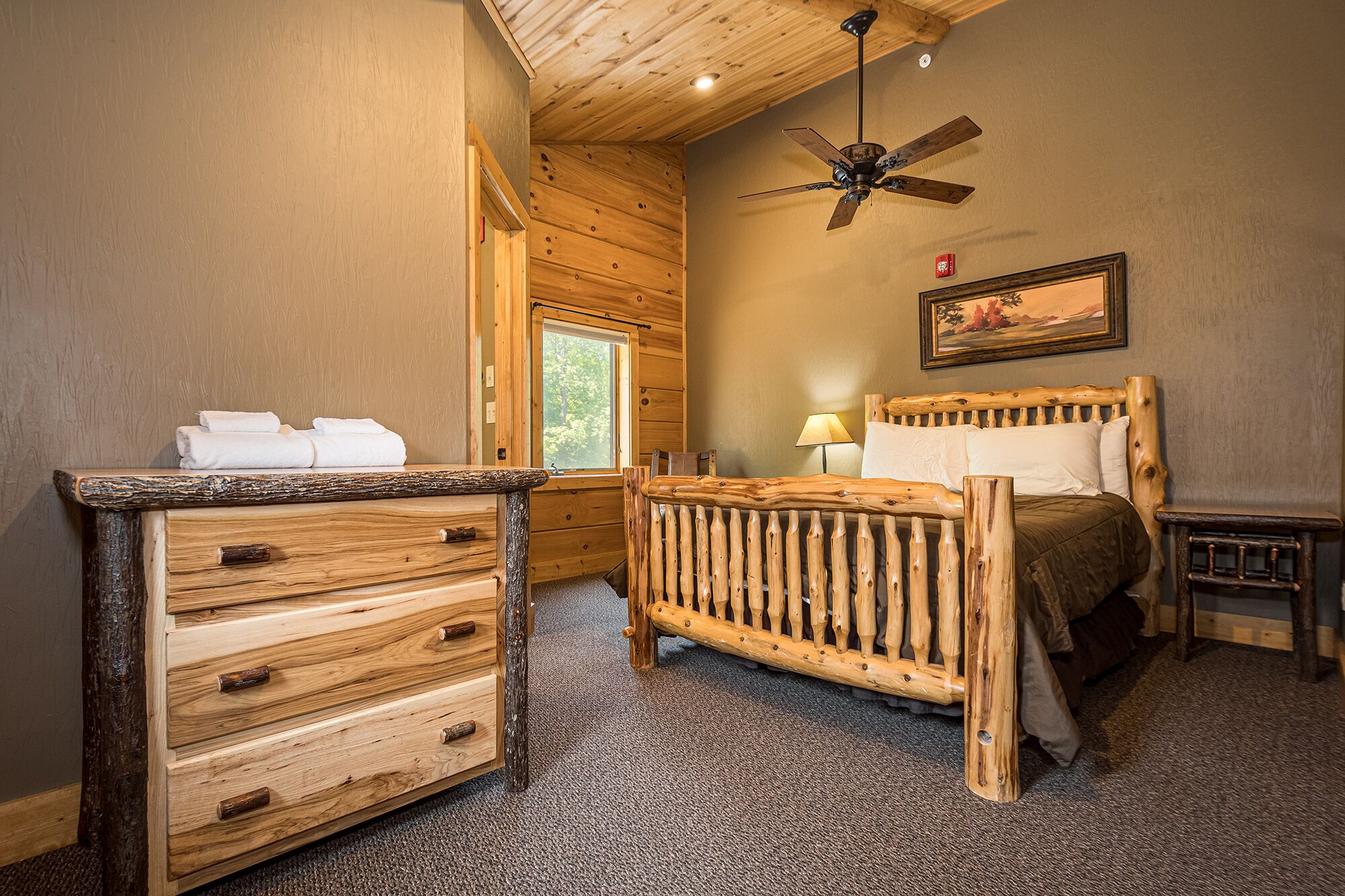 Un dormitorio rústico de madera con cama, cómoda de madera, ventilador de techo y ventana.