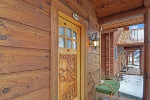 A wooden cabin with a carved door, a porch with two chairs, and a view of a snowy landscape. - C46R - 3 Queen Beds, Duplex (McHenry)