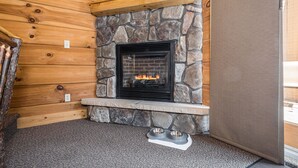 A stone fireplace with a fire, set in a log cabin-style room with a stone accent wall and a view of a forest landscape. - Mountainside Loft Cabin 46R (McHenry)
