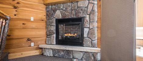 Une cheminée en pierre avec un feu, située dans une pièce aménagée dans le style d’un chalet en rondins, avec un mur d’accent en pierre et une vue sur un paysage forestier.