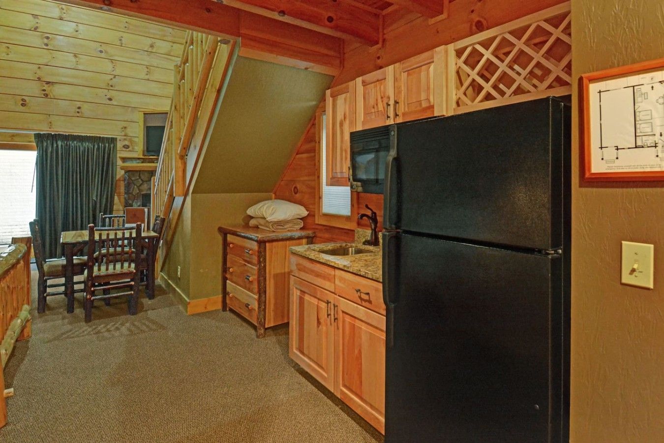 Une cuisine avec des meubles de rangement en bois, un réfrigérateur noir, une table à manger avec des chaises et un escalier menant à l’étage supérieur.