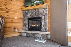 A stone fireplace with a fire, set in a wooden cabin with a stone hearth and a mounted television above it. - C46R - 3 Queen Beds, Duplex (McHenry)