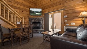 A cozy cabin interior with a stone fireplace, wooden walls, a television mounted above it, and a leather couch. - Mountainside Loft Cabin 46R (McHenry)