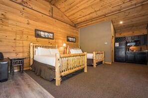 A log cabin-style room with two wooden beds, a small table, and a kitchenette with dark cabinets.