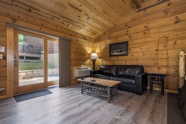 A wooden interior with a sliding glass door leading outside, a black leather sofa, a small wooden table, and a lamp.