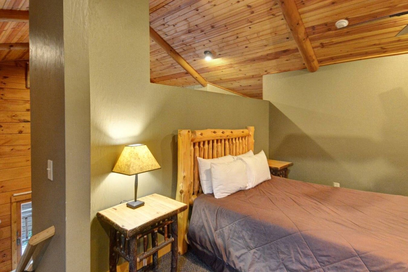 A bedroom with a wooden ceiling, a bed with a headboard, a nightstand, and a lamp.