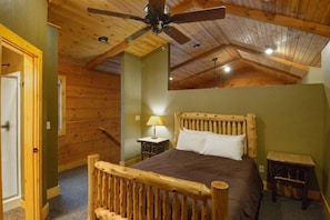 Un dormitorio con ventilador de techo de madera, cama con cabecera de madera, mesita de noche con lámpara y un taburete pequeño de madera.