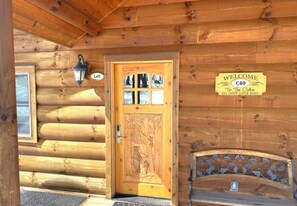A wooden cabin entrance with a carved door, a welcome sign, and a bench outside. - C49L - 2 Queen Beds, Duplex (McHenry)