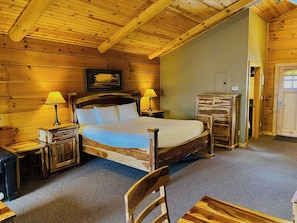A wooden cabin bedroom with a bed, two bedside tables, a chair, and a door. - C49R - King Bed, Duplex (McHenry)