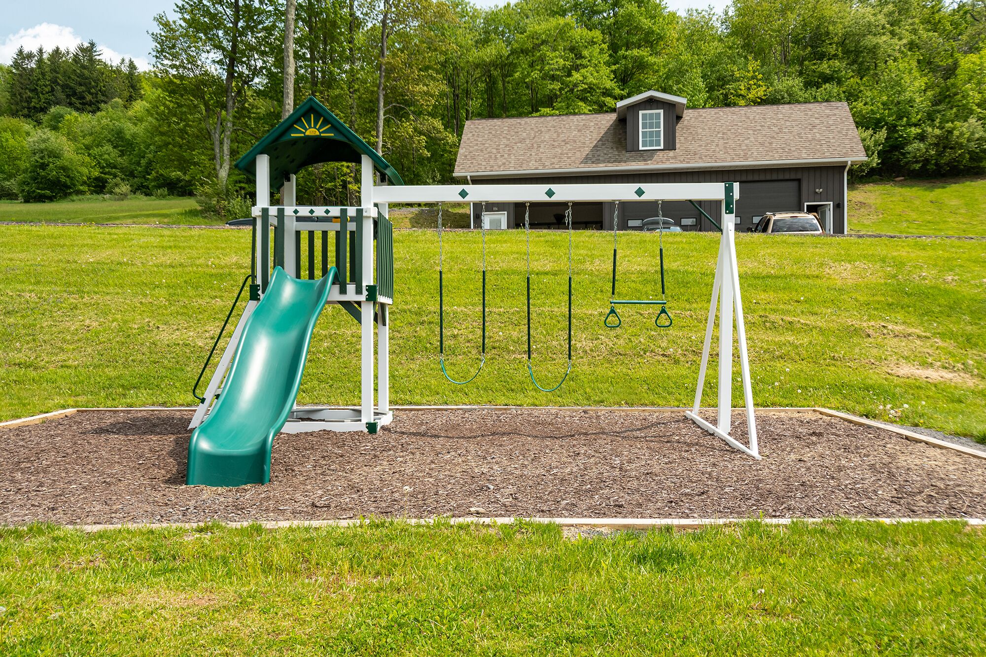 A playground with a slide and swings, surrounded by a grassy field and a building in the background.