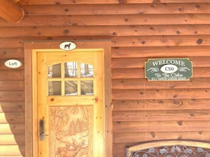 Entrada de una cabaña de madera con una puerta decorada con una talla de ciervo, un cartel de bienvenida y una señalización que indica la entrada a la izquierda.