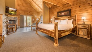A log cabin bedroom with a stone fireplace, a bed with a wooden headboard, a desk with a chair, and a television mounted on the wall.
