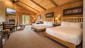 A log cabin room with two beds, a stone fireplace, a TV, and a balcony.