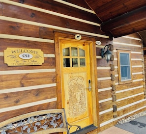 Una cabaña de madera con puerta tallada, un letrero de bienvenida y un banco.