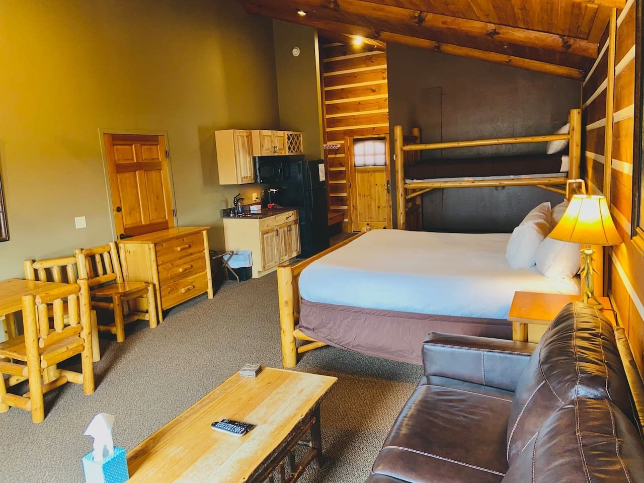 A compact room with a bunk bed, wooden furniture, a small kitchenette, and a single bed with a bedside table.