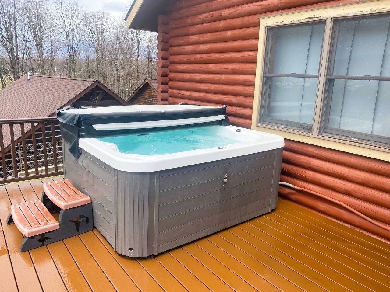 Un jacuzzi en una terraza de madera con escalones que conducen a él, rodeado por una estructura tipo cabaña de madera.