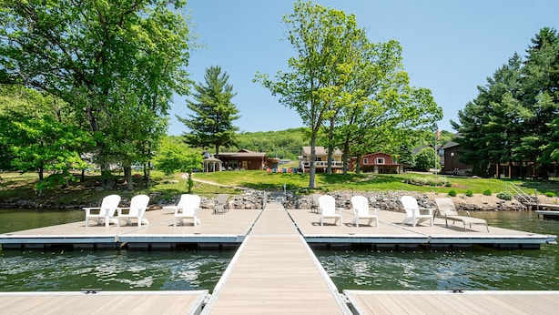 Een houten steiger met witte Adirondack-stoelen die uitkomt bij een kalm meer, omringd door weelderige begroeiing en een klein vakantiehuisje op de achtergrond.