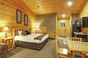 A log cabin-style room with a bed, bedside lamps, a wooden table, and a kitchen area.
