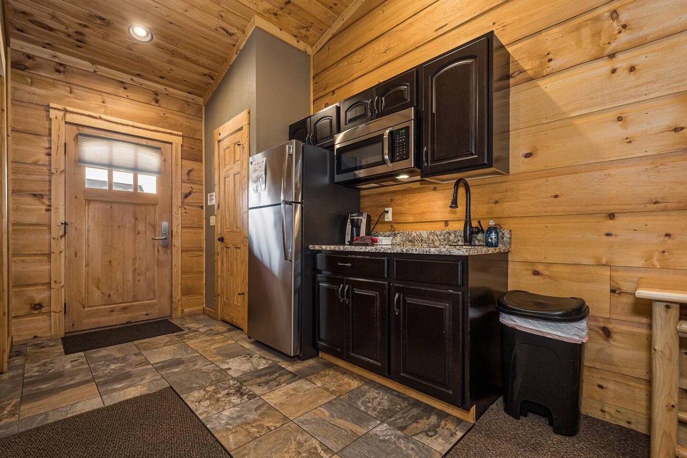 A compact kitchen with wooden walls, a refrigerator, a microwave, and a sink.