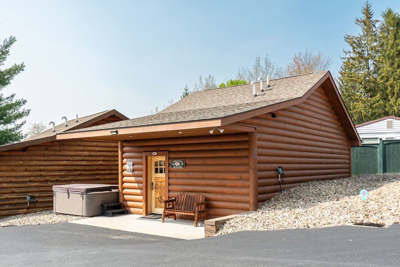 A log cabin with a wooden door, a bench, and a covered area with a trash bin.