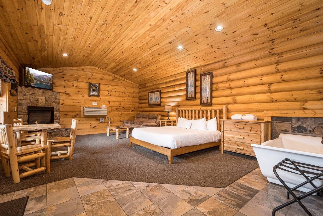 A log cabin interior with a stone fireplace, a bed, a desk, and a bathtub.