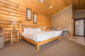 Ein Schlafzimmer in einer Blockhütte mit einem hölzernen Bett, einem Nachttisch, einer Lampe, einem Schminktisch und einem Badezimmer mit Badewanne und Dusche.