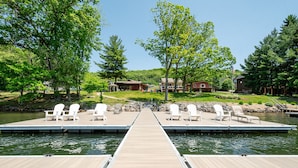 Une jetée en bois, agrémentée de chaises Adirondack blanches, mène à un lac paisible, entouré d’une végétation luxuriante et surmonté, au loin, d’une petite cabane.