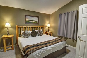 Une chambre à coucher avec une tête de lit en bois, un lit recouvert d’une literie à motifs, deux tables de chevet avec des lampes, un tableau accroché au mur et une porte ornée de rideaux.