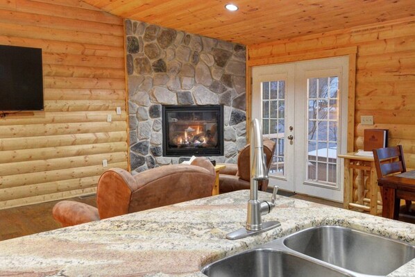A log cabin interior with a stone fireplace, a stone wall, a wooden ceiling, a stone countertop with a sink, and a television mounted on the wall.