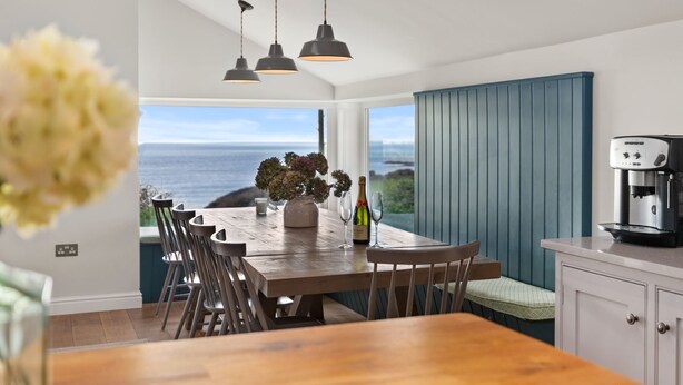 Un coin repas avec une table en bois, des chaises et une vue sur la mer. Trois lampes suspendues sont placées au-dessus de la table, et une machine à café est visible sur la droite.