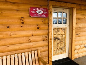 Una cabaña de madera con un letrero de bienvenida, un banco y una puerta decorada con un grabado de pez.