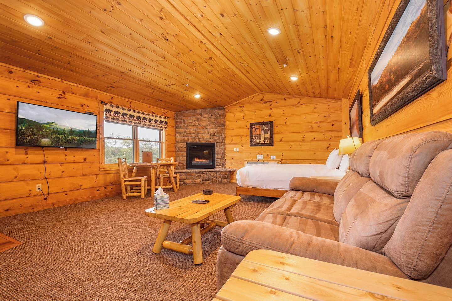 A cozy cabin interior with a stone fireplace, wooden walls, a large window, and a television.