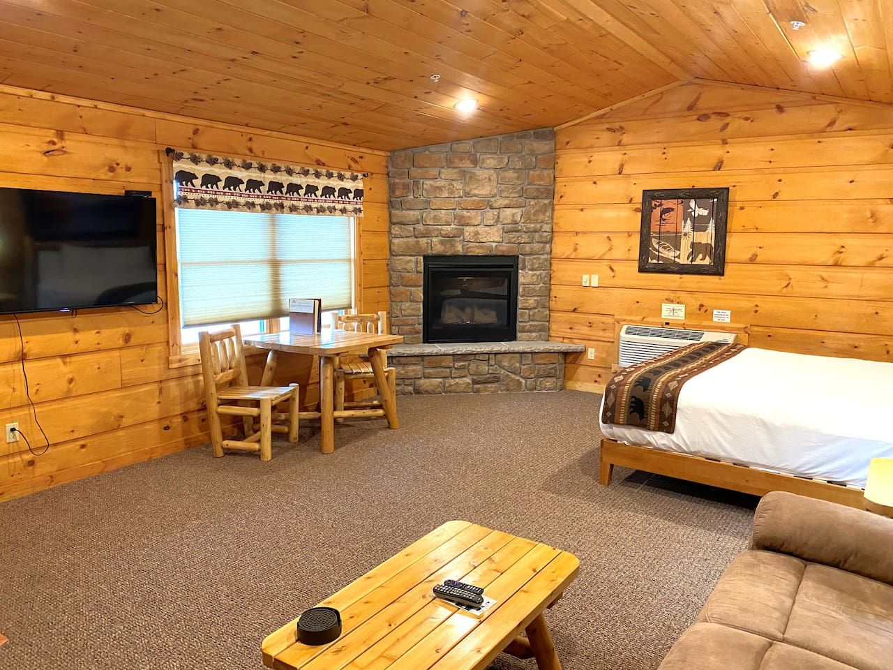 A cabin interior with a stone fireplace, wooden furniture, a bed, and a television.