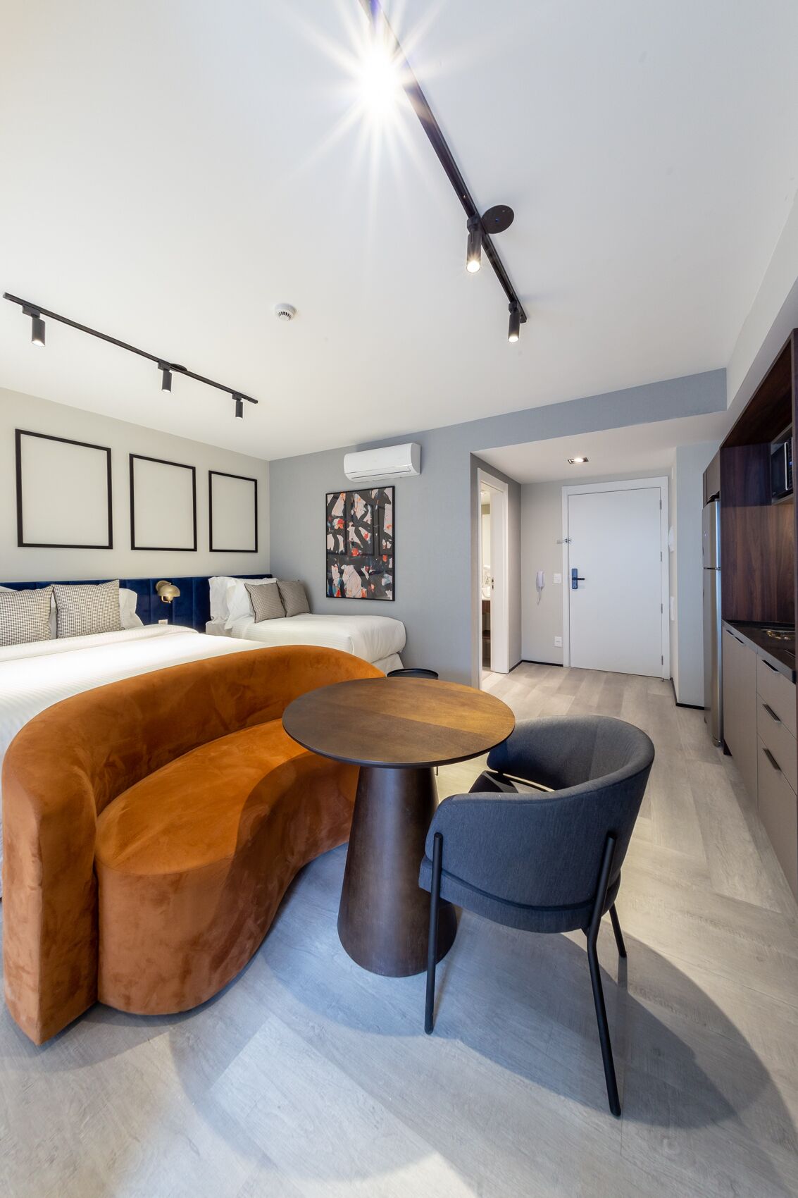 A modern living room with a curved sofa, a round coffee table, and a blue chair. There is a bed with white bedding and a painting on the wall.