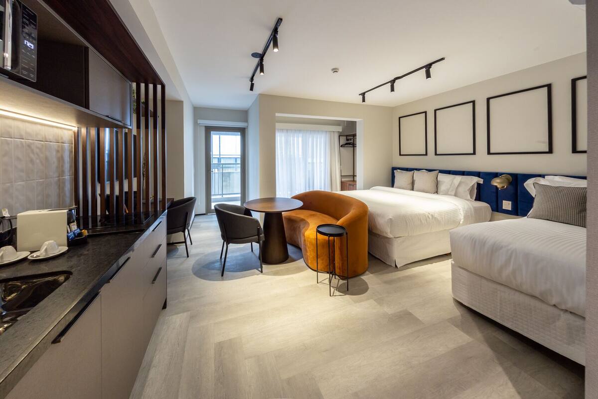 A modern hotel room with a kitchen area, a dining table with chairs, a sofa, a bed, and a balcony with a view.