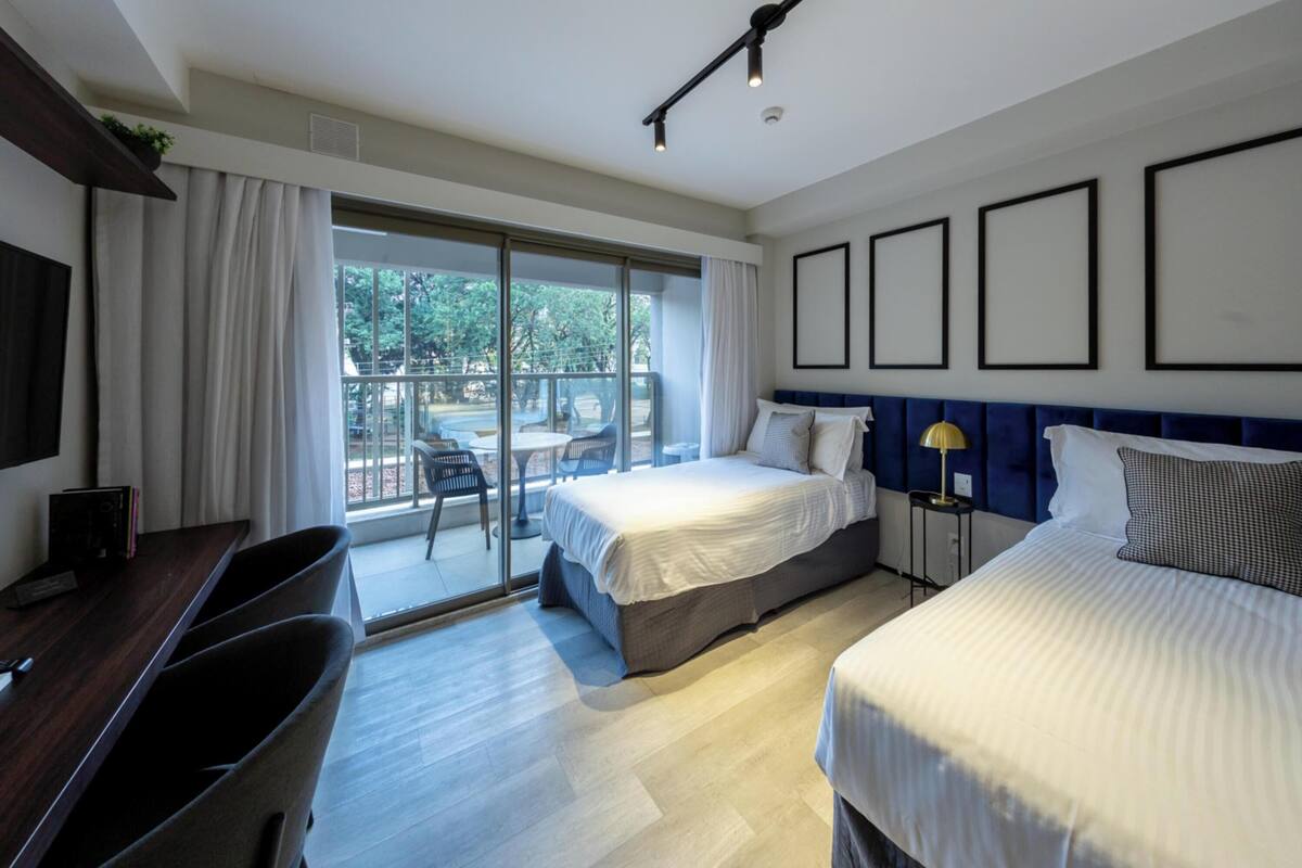 A modern hotel room with a large bed, a desk, and a balcony with outdoor seating.
