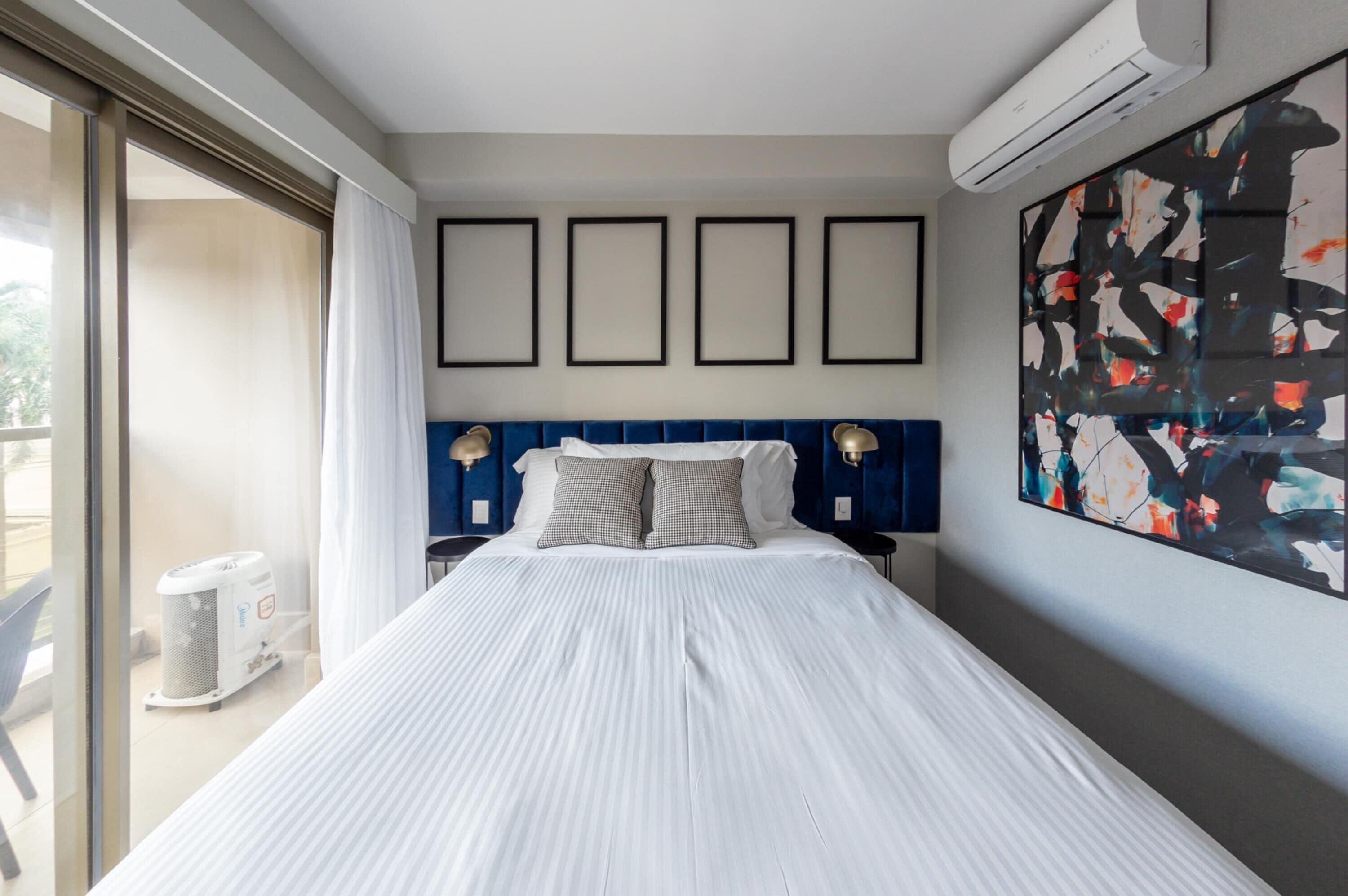 A hotel room with a large bed, a blue headboard, a wall-mounted air conditioner, and a large abstract painting.