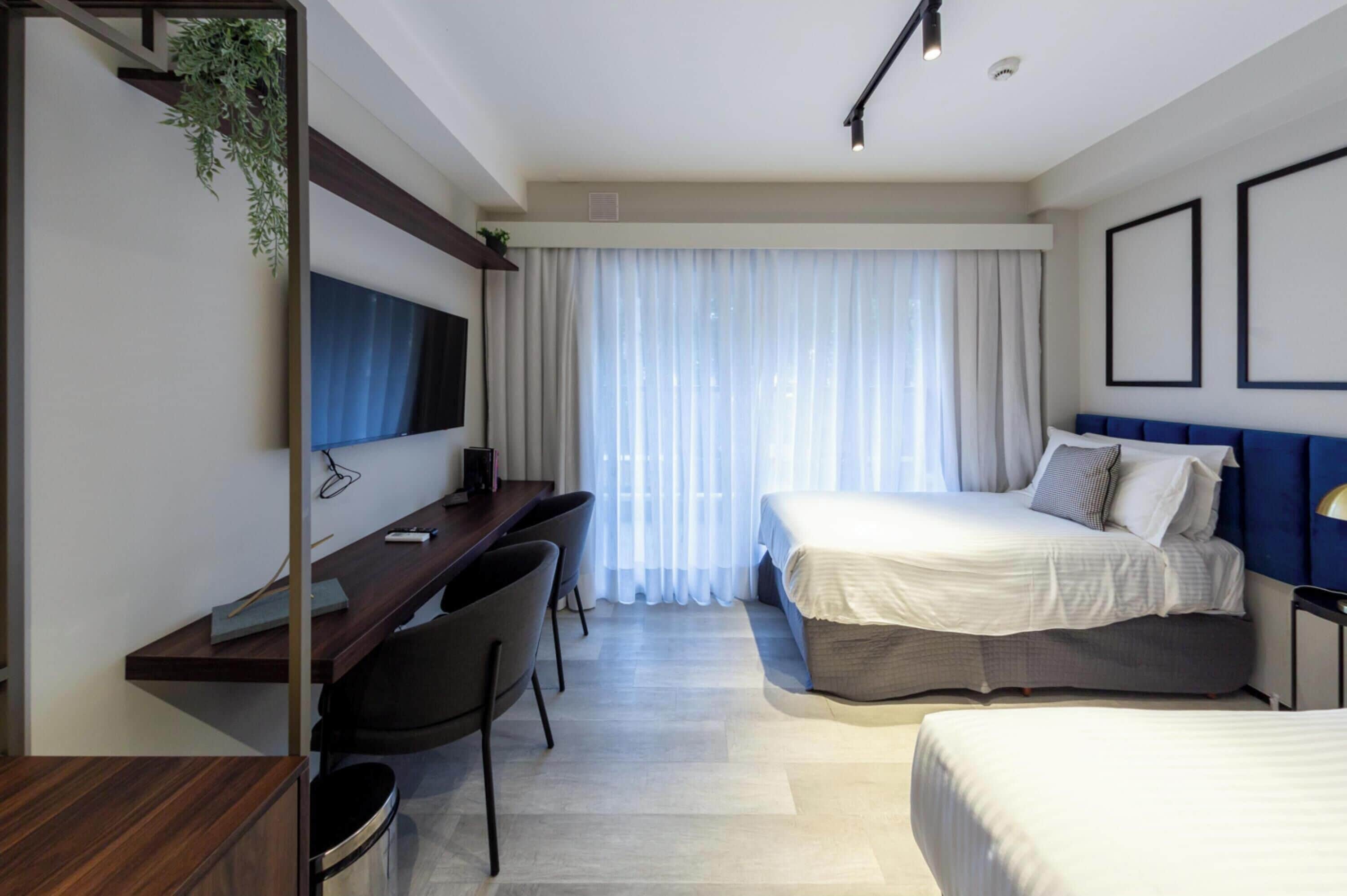 A hotel room with a desk, two beds, a TV, and a large window with curtains.