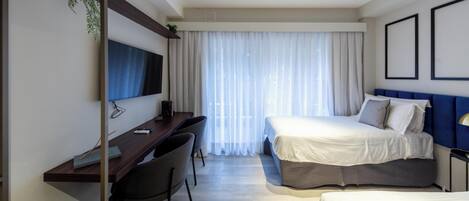 A hotel room with a desk, two beds, a TV, and a large window with curtains.