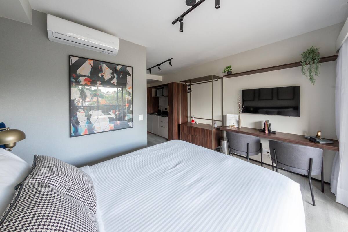 A modern hotel room with a large bed, a desk with two chairs, a mounted TV, and a kitchen area in the background.