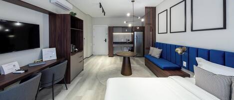 A modern hotel room with a large bed, a desk with a flat-screen TV, a seating area with a blue sofa, and a kitchenette in the background.