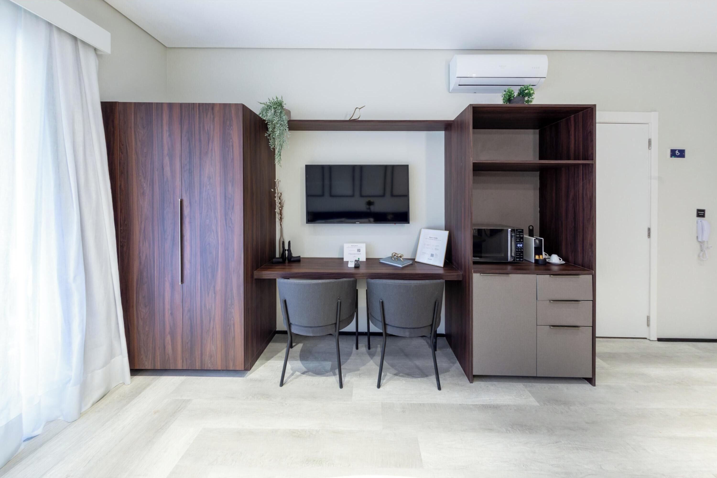 Quarto de hotel moderno com escrivaninha de madeira, duas cadeiras, televisão de tela plana, micro-ondas e uma pequena planta.