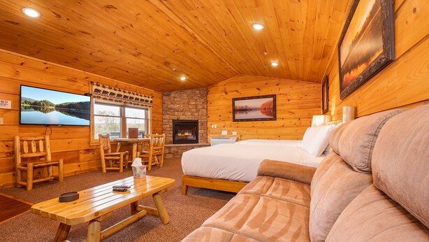A cozy cabin interior with a wooden ceiling, a stone fireplace, a bed, a sofa, and a television on the wall. - Grandview Cabin Suite 24-Hot Tub (McHenry)