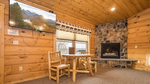 A wooden cabin interior with a stone fireplace, a dining table with chairs, a television mounted on the wall, and a scenic view of mountains and a lake through the window. - Grandview Cabin Suite 27-Hot Tub, Dog Friendly (McHenry)