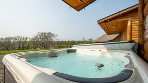 Un jacuzzi con terraza de madera, una cabaña de madera al fondo y cielo despejado.